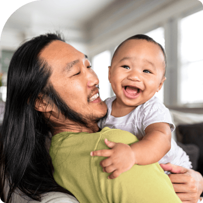 Asian man with long hair wearing a lime green shirt holding a baby they are both smiling at the camer Asian man with long hair wearing a lime green shirt holding a baby they are both smiling at the camer