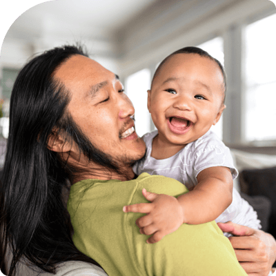 Asian man with long hair wearing a lime green shirt holding a baby they are both smiling at the camer